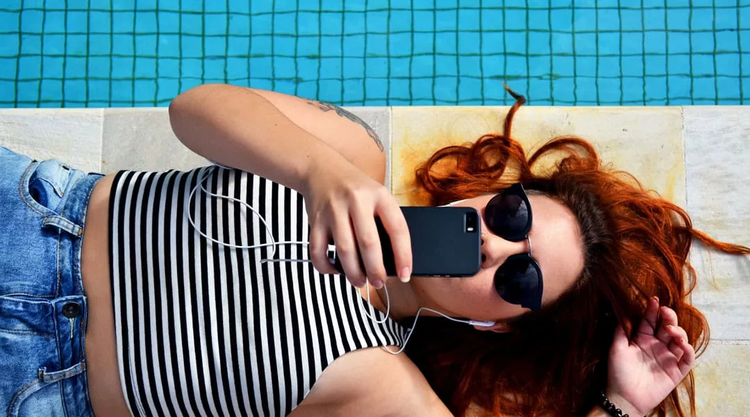 woman with red hair in jeans and stripped shirt lying poolside with headphone on using her phone to create and invoice using Joy Pilots AI invoicing feature