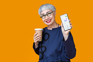 older woman with short haircut and wearing glasses holding a coffee in one hand while presenting to you the screen of her phone showing the invoices sales page of her company's online accounting account on joy pilot